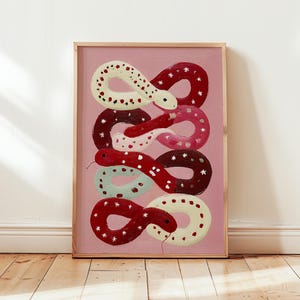 Op de afbeelding: Een ingelijste kunstdruk met een gestileerde illustratie van slangen in een herhalend patroon. De slangen zijn in tinten rood, wit, roze en blauwgroen, met stervormige details, tegen een roze achtergrond.