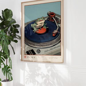 May include: A framed art print featuring a green frog on a blue record on a turntable. The frog is perched on a red center label. The turntable has a silver body and a red tone arm. The background is light blue.