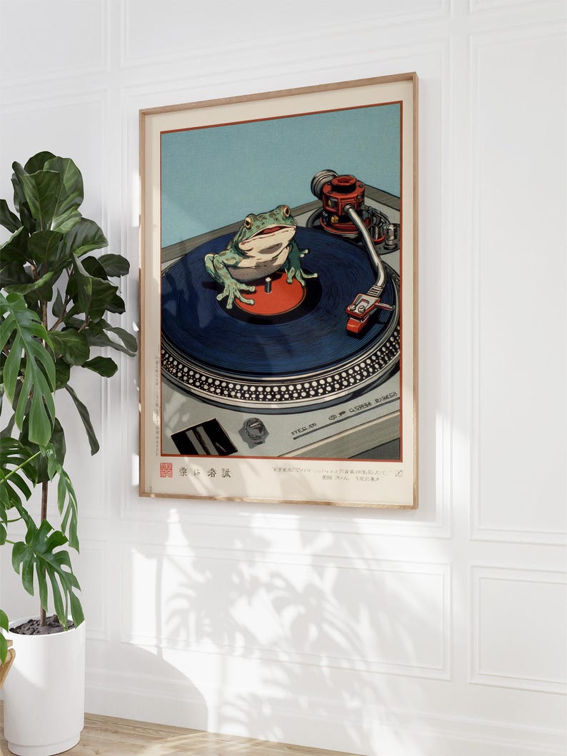 May include: A framed art print featuring a green frog on a blue record on a turntable. The frog is perched on a red center label. The turntable has a silver body and a red tone arm. The background is light blue.