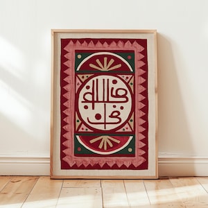 May include: Framed artwork featuring Arabic calligraphy in a circular design, set against a red and green geometric background. The artwork is framed in a light wood frame and is displayed against a white wall.
