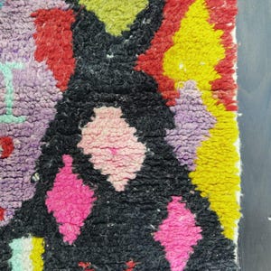 May include: Close-up of a colorful, textured rug with a geometric design. The rug features a black central shape with pink diamond patterns, surrounded by red, yellow, green, purple, and blue accents. The rug has a thick, woven texture.