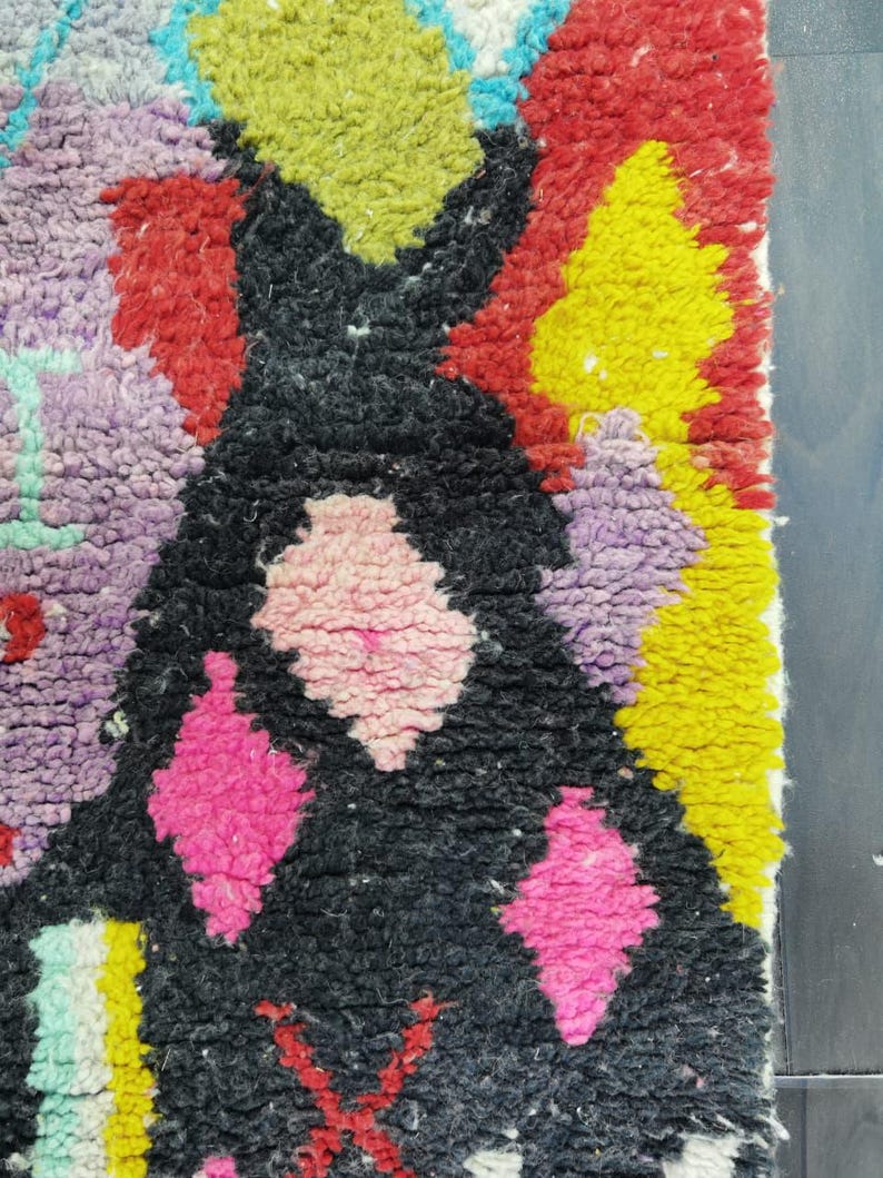 May include: Close-up of a colorful, textured rug with a geometric design. The rug features a black central shape with pink diamond patterns, surrounded by red, yellow, green, purple, and blue accents. The rug has a thick, woven texture.