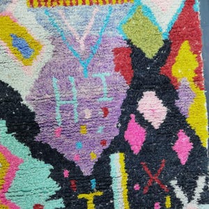 May include: Close-up of a colorful, textured rug with a geometric design. The rug features a mix of vibrant colors including purple, black, pink, yellow, and turquoise. The design includes diamond shapes and abstract patterns.