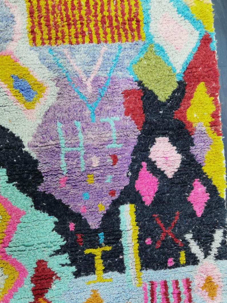 May include: Close-up of a colorful, textured rug with a geometric design. The rug features a mix of vibrant colors including purple, black, pink, yellow, and turquoise. The design includes diamond shapes and abstract patterns.