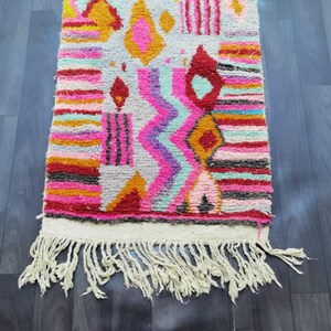 May include: A colorful, rectangular rug with a geometric pattern. The rug features a variety of colors, including pink, orange, yellow, and blue. It has a white fringe along one edge. The rug is made of a thick, woven material.