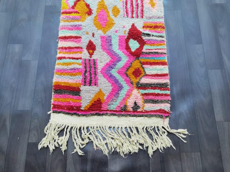 May include: A colorful, rectangular rug with a geometric pattern. The rug features a variety of colors, including pink, orange, yellow, and blue. It has a white fringe along one edge. The rug is made of a thick, woven material.