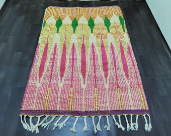May include: A rectangular rug with a colorful geometric pattern. The rug features a cream-colored background with vertical pink and yellow stripes. Green diamond shapes and orange lines are at the top. The rug has a purple border and white tassels.