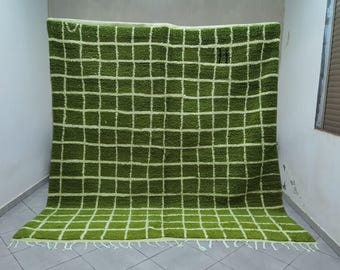 May include: A large, green rug with a grid pattern of white lines. The rug is made of a thick, textured material and has a fringe along the bottom edge. The rug is displayed against a white wall.