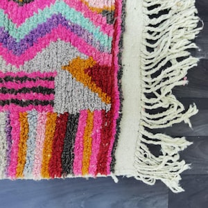 May include: Close-up of a colorful rug with a geometric pattern. The rug features a vibrant mix of pink, orange, red, purple, and teal hues. The rug has a white fringe along one edge, adding texture and detail to the design.