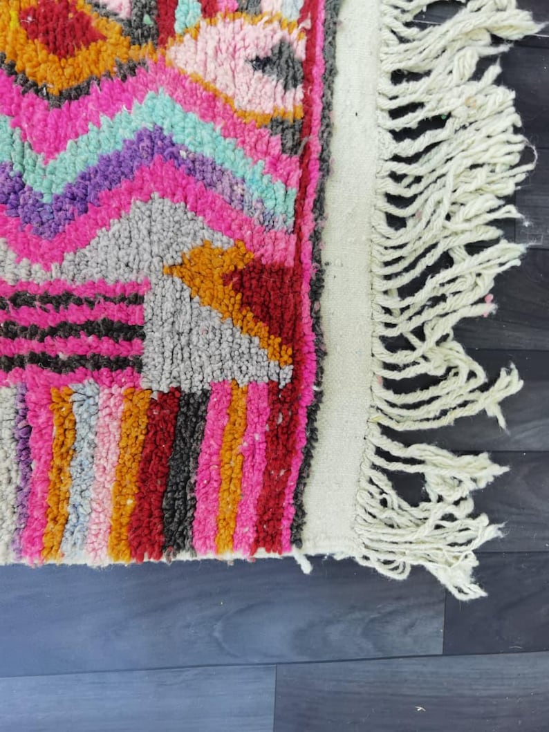 May include: Close-up of a colorful rug with a geometric pattern. The rug features a vibrant mix of pink, orange, red, purple, and teal hues. The rug has a white fringe along one edge, adding texture and detail to the design.
