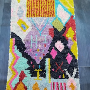 May include: A colorful, patterned rug with a geometric design. The rug features a variety of shapes and colors, including yellow, pink, black, and purple. The rug is made of a soft, textured material and is likely used as a decorative floor covering.