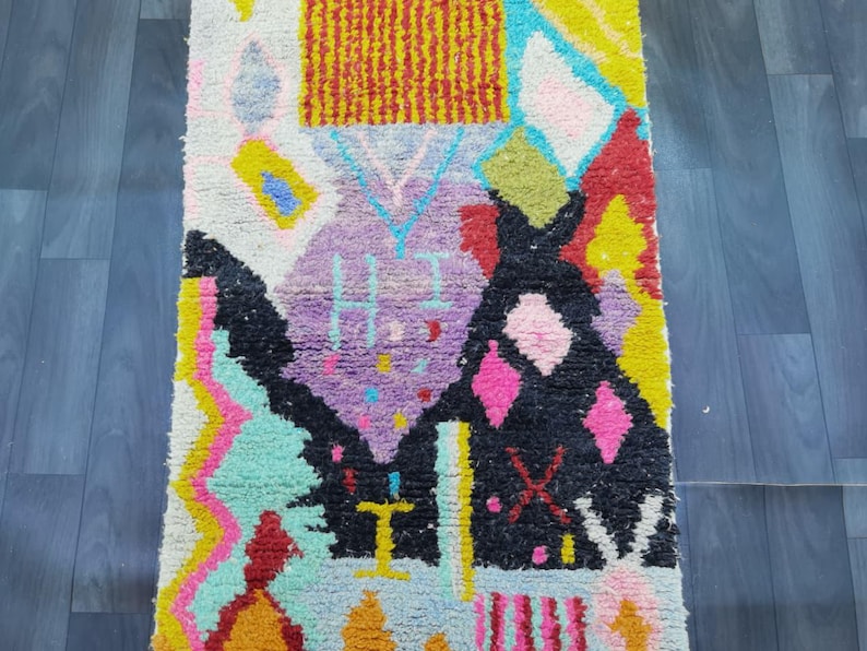 May include: A colorful, patterned rug with a geometric design. The rug features a variety of shapes and colors, including yellow, pink, black, and purple. The rug is made of a soft, textured material and is likely used as a decorative floor covering.