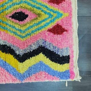 May include: Close-up of a pink, geometric patterned rug. The rug features a diamond pattern in yellow, blue, and black, with red diamond accents. The rug has a textured, woven appearance, with a white border.