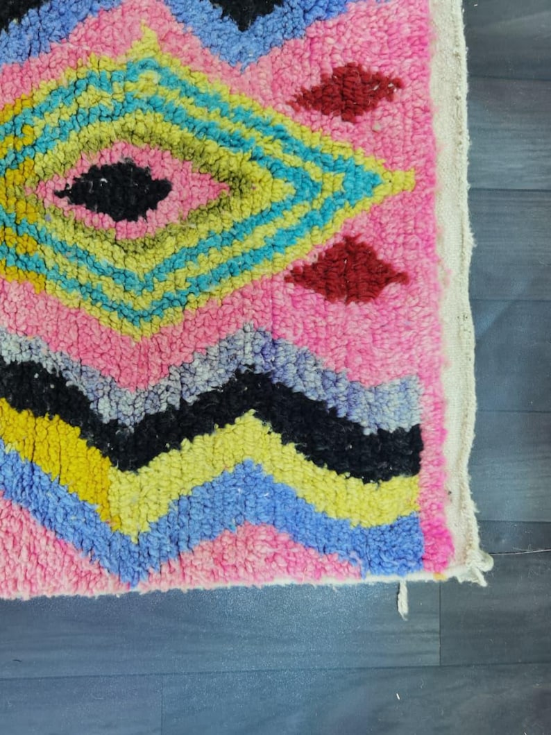May include: Close-up of a pink, geometric patterned rug. The rug features a diamond pattern in yellow, blue, and black, with red diamond accents. The rug has a textured, woven appearance, with a white border.