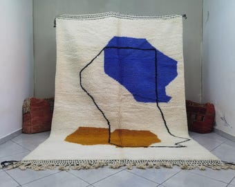 May include: A white wool rug with a blue and brown abstract design. The rug has a fringe border.