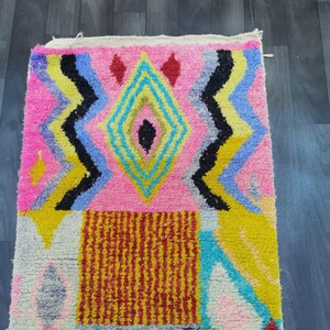 May include: A colorful, rectangular rug with a geometric design. The rug features a pink background with a central diamond shape in yellow, blue, and black. Additional patterns include zigzags and a red and yellow rectangle.