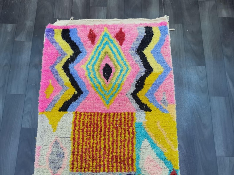 May include: A colorful, rectangular rug with a geometric design. The rug features a pink background with a central diamond shape in yellow, blue, and black. Additional patterns include zigzags and a red and yellow rectangle.