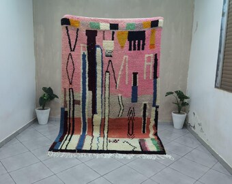Op de afbeelding: Een roze tapijt met abstracte patronen met een verscheidenheid aan vormen en kleuren. Het tapijt heeft verticale en horizontale lijnen, ruiten en andere geometrische vormen in tinten roze, blauw, zwart en wit. Het tapijt wordt tegen een neutrale muur getoond.