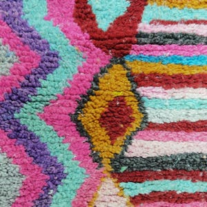 May include: Close-up of a colorful, textured rug with a geometric pattern. The rug features a vibrant mix of colors, including pink, purple, turquoise, red, yellow, and black. The design includes diamond and zigzag shapes.