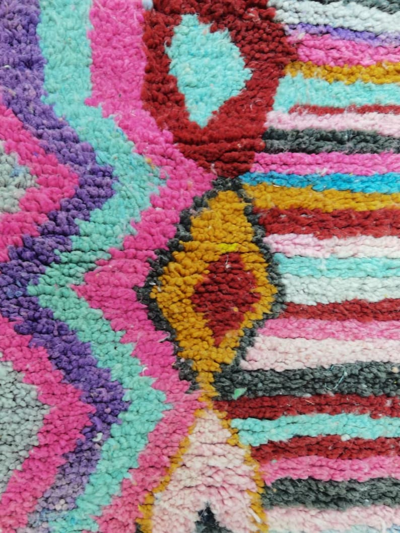 May include: Close-up of a colorful, textured rug with a geometric pattern. The rug features a vibrant mix of colors, including pink, purple, turquoise, red, yellow, and black. The design includes diamond and zigzag shapes.