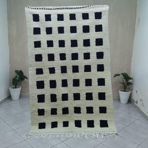 May include: A rectangular, cream-colored rug with a repeating pattern of black squares. The rug has a fringe along the bottom edge. The rug is displayed in a room with white walls and tile flooring. The rug is approximately 5 feet wide and 7 feet long.