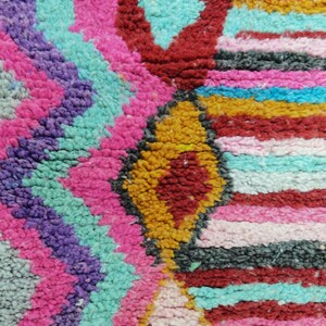 May include: Close-up of a colorful, textured rug with a geometric pattern. The rug features a vibrant mix of colors, including pink, purple, turquoise, red, yellow, and black. The design includes diamond and zigzag shapes.