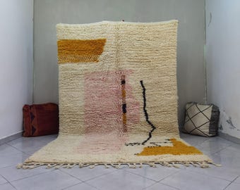 May include: Cream-colored, textured rug with abstract design elements. Features a large mustard-yellow rectangle, a pink vertical stripe, and a black curved line. The rug has a fringe along the bottom edge. Decorative floor covering.