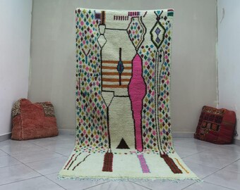 May include: A rectangular, cream-colored rug with a geometric design. The rug features a central pink vertical stripe, brown and blue horizontal stripes, and colorful diamond shapes. The rug is approximately 6 feet wide and 9 feet long.