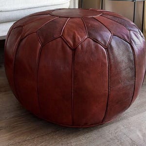 Könnte beinhalten: Ein runder, dunkelroter Lederhocker mit geometrischem Muster. Der Hocker hat einen tiefen Burgunderfarbton und scheint aus genähten Lederpaneelen gefertigt zu sein. Es ist ein niedriges, bodennahes Möbelstück.