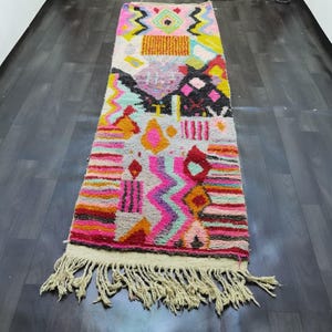 May include: A rectangular, colorful rug with a cream fringe. The rug features a geometric design with pink, orange, yellow, black, and turquoise shapes. The rug is placed on a dark wood floor.