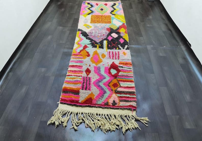 May include: A rectangular, colorful rug with a cream fringe. The rug features a geometric design with pink, orange, yellow, black, and turquoise shapes. The rug is placed on a dark wood floor.