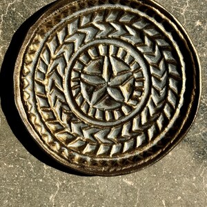 Peut inclure: Une assiette ronde en céramique avec un motif d'étoile au centre. L'assiette présente un motif en relief texturé de chevrons et d'une étoile. Elle est vernie dans des tons bronze et gris.
