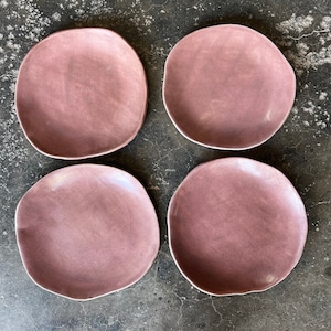 May include: Four dusty rose colored ceramic plates with a slightly irregular shape. The plates are arranged in two rows of two on a gray concrete surface.