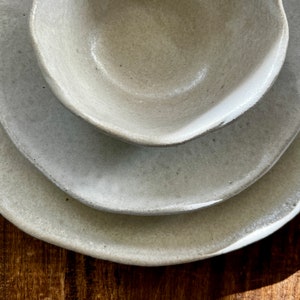 May include: A set of three ceramic bowls in a light gray color. The bowls are nested together, with the smallest bowl on top. The bowls have a slightly textured surface and a rounded shape.