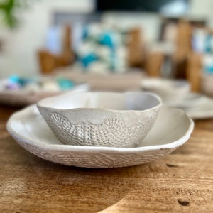May include: A white ceramic bowl with a decorative lace pattern sits inside a larger white ceramic bowl with a similar pattern. Both bowls are on a wooden surface.