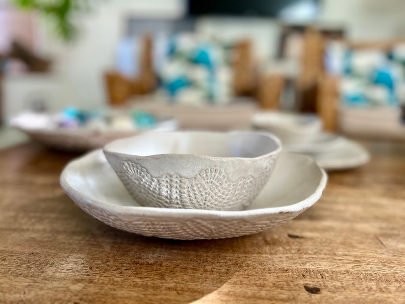 May include: A white ceramic bowl with a decorative lace pattern sits inside a larger white ceramic bowl with a similar pattern. Both bowls are on a wooden surface.
