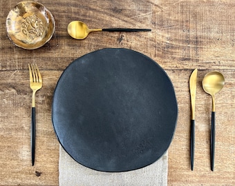 May include: A black matte dinner plate with gold and black flatware, including a fork, knife, and spoon. A small, decorative gold dish is at the top left. The items are arranged on a wooden surface.