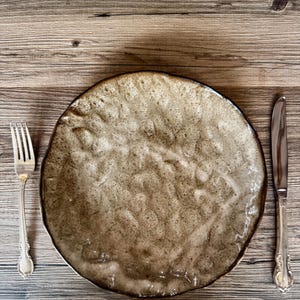 May include: A rustic, handmade ceramic plate with a textured, mottled brown surface. A silver fork and knife are placed on either side of the plate, set against a wooden background. The plate has a unique, organic shape.