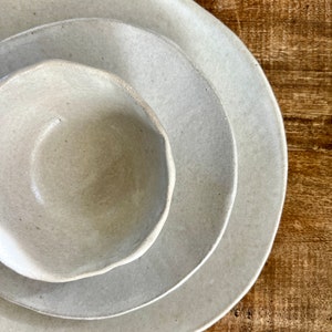 May include: Three ceramic bowls in various sizes, all in a light beige color, with a slightly textured surface. The bowls are stacked on top of each other, with the largest bowl on the bottom and the smallest bowl on top.