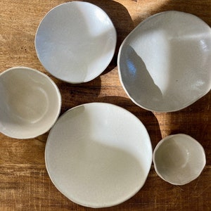 May include: Five ceramic bowls in various sizes and shapes, all in a light gray color. The bowls are arranged on a wooden surface.