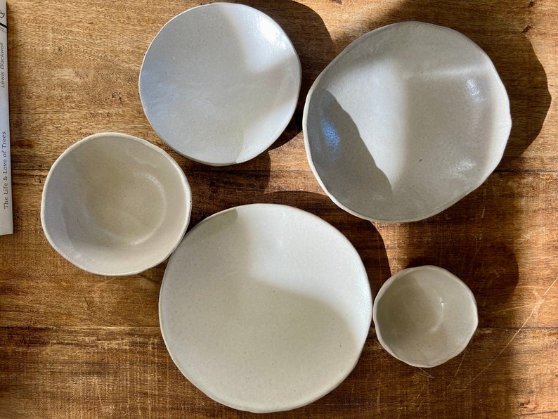 May include: Five ceramic bowls in various sizes and shapes, all in a light gray color. The bowls are arranged on a wooden surface.