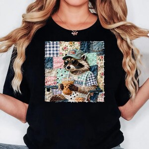 May include: Black t-shirt with a raccoon wearing sunglasses and a cap, holding a drink with a straw. The raccoon is set against a patchwork background with floral and plaid patterns. The shirt has the word "IBMAN" on it.