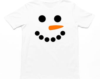 May include: White t-shirt featuring a snowman face design. The design includes two large black circles for eyes, an orange carrot nose, and a row of seven black circles for the mouth, creating a cheerful winter theme.