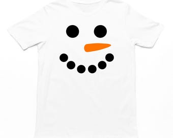 May include: White t-shirt featuring a snowman face design. The design includes two large black circles for eyes, an orange carrot nose, and a smiling mouth made of seven small black circles. A festive and fun winter-themed apparel item.