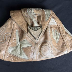 May include: A tan handbag featuring a textured, reptilian-patterned surface with contrasting cream-colored, perforated leather appliques in abstract shapes. The bag has a triangular front flap secured by a metallic, V-shaped hardware accent.