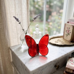 May include: A vibrant red glass butterfly figurine with translucent wings and a dark red central body. The butterfly is crafted from smooth, polished glass, featuring symmetrical wing shapes that catch the light, creating a clear and bold visual statement.