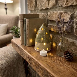 May include: A cone-shaped ceramic luminary with a green glaze and circular cutouts, illuminated from within. It sits beside three vintage books, a small clear glass vase containing dried lavender sprigs, and a single brown pinecone.