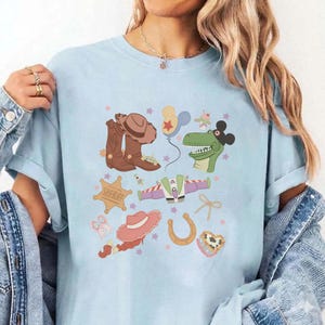 May include: Light blue t-shirt featuring a colorful graphic of cowboy boots, a sheriff's badge, a cowboy hat, a horseshoe, and a dinosaur. The design also includes a space ship, balloons, and stars.