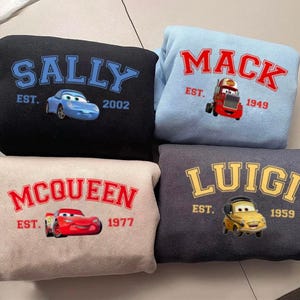 May include: Four sweatshirts featuring cartoon car designs. The black sweatshirt has "SALLY" in blue, a blue car graphic, and "EST. 2002". The light blue sweatshirt says "MACK" in red, a red truck graphic, and "EST. 1949". The beige sweatshirt says "MCQUEEN" in red, a red car graphic, and "EST. 1977". The dark grey sweatshirt says "LUIGI" in yellow, a yellow car graphic, and "EST. 1959".