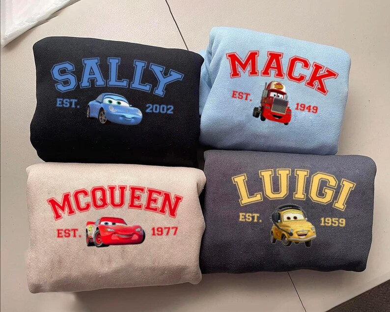 May include: Four sweatshirts featuring cartoon car designs. The black sweatshirt has "SALLY" in blue, a blue car graphic, and "EST. 2002". The light blue sweatshirt says "MACK" in red, a red truck graphic, and "EST. 1949". The beige sweatshirt says "MCQUEEN" in red, a red car graphic, and "EST. 1977". The dark grey sweatshirt says "LUIGI" in yellow, a yellow car graphic, and "EST. 1959".